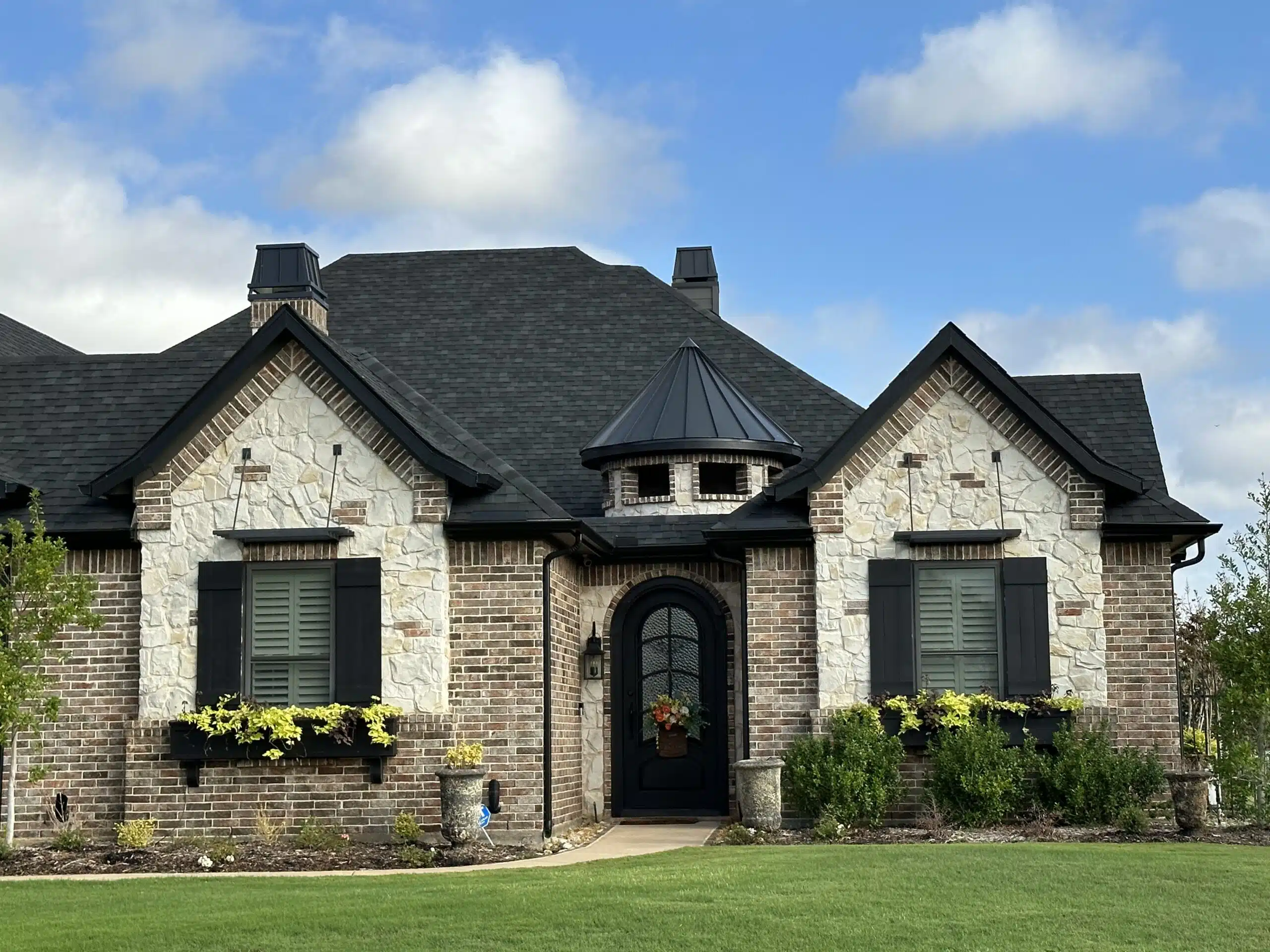 A two-story brick house with stone accents, black shutters, and a dark grey roof. The arched front door is flanked by two flower pots, and a small garden area with shrubs and flowers surrounds the entrance. The sky is blue with scattered clouds.