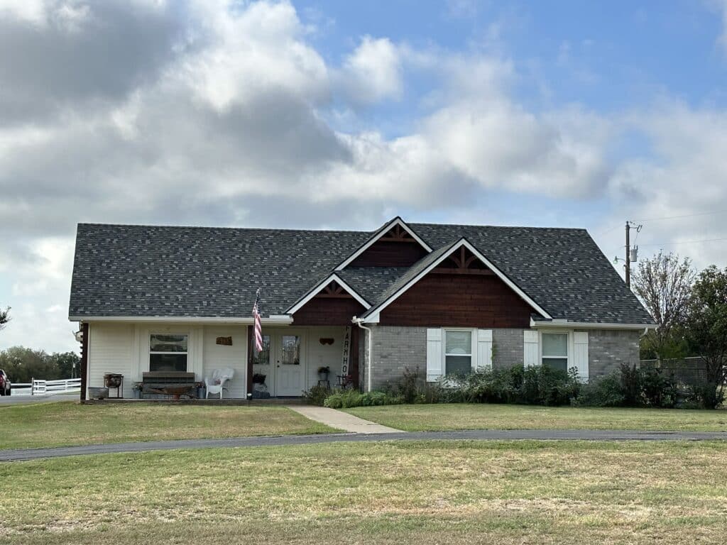 A single-story house with a grey shingle roof, white and grey brick exterior, and two small dormer windows. An American flag hangs near the front porch, and there are chairs and potted plants outside. The house is surrounded by grass and a few trees, under a partly cloudy sky.