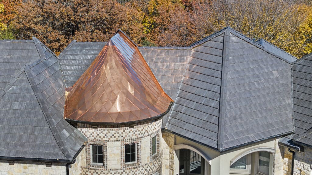 A rooftop view of a house with a unique, conical copper roof segment surrounded by black slate roofing. The building features stone walls with decorative brick patterns. In the background, there are trees with vibrant autumn foliage in shades of orange, yellow, and red.