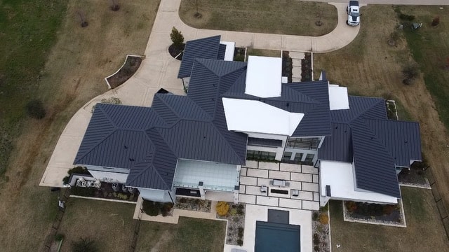 alt= Aerial view of a large modern house with multiple metal roofs, surrounded by a landscaped yard and a pool, with a driveway leading to the street.