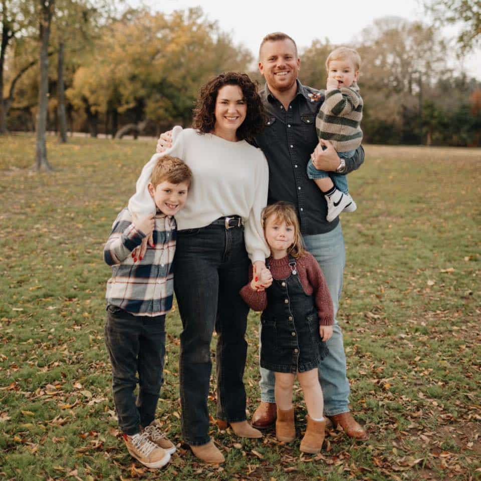 A family of five stands outdoors on a grassy field with trees in the background. The parents stand in the middle, each holding one child's hand. The father holds a baby, while the mother stands beside an older boy and girl. They all are smiling and wearing casual clothing.