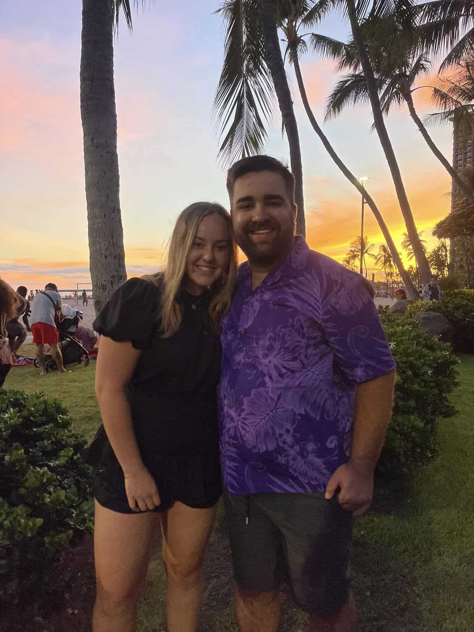 A smiling man and woman stand close together outdoors with palm trees and a sunset in the background. The man is wearing a purple shirt, and the woman is wearing a black dress. Other people can be seen in the distance near the beach.