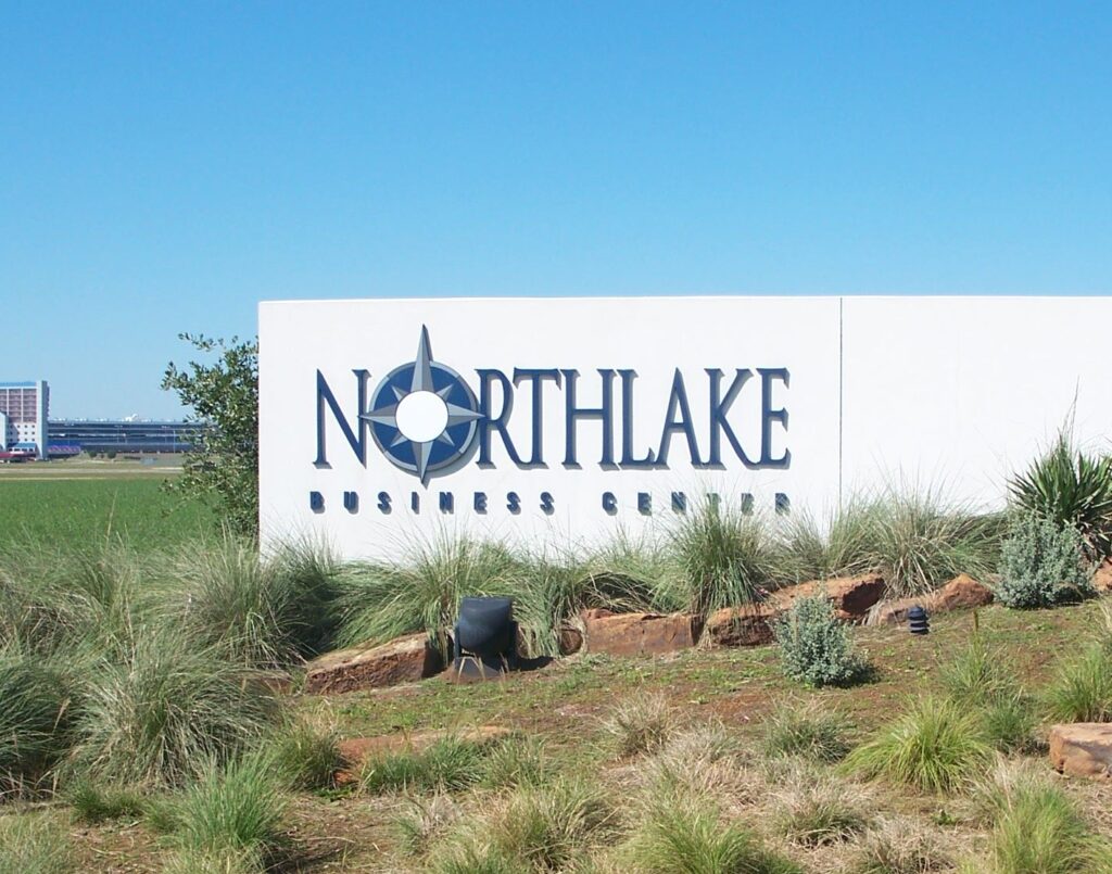 A large white sign reads "Northlake Business Center" with a stylized compass design integrated into the text. The sign is surrounded by landscaped vegetation, including grasses and small shrubs. A clear blue sky is visible in the background.