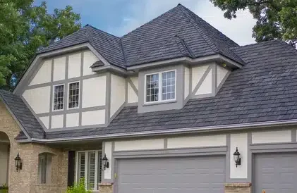 A well-maintained two-story house with a Tudor-style exterior, featuring white walls, grey trim, and a dark grey, steeply pitched roof. The front yard includes blooming yellow flowers and a paved driveway leading to a two-car garage. Trees surround the property.