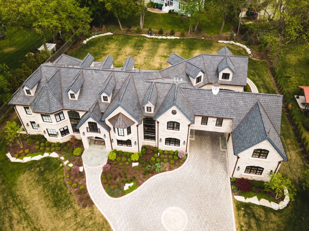 Aerial view of a large, modern, two-story mansion with multiple peaked roofs and large windows. The house is surrounded by a well-manicured lawn with flower beds and stone pathways. An expansive driveway leads to a garage connected to the main building.