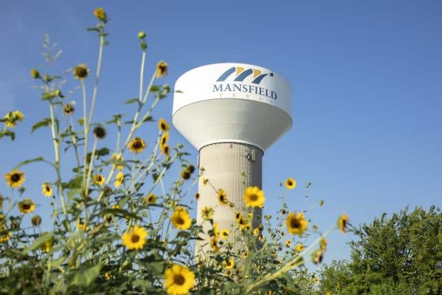 A tall water tower with "Mansfield, Texas" written on it stands against a clear blue sky. Yellow flowers in the foreground and green trees in the background add a touch of nature to the scene.