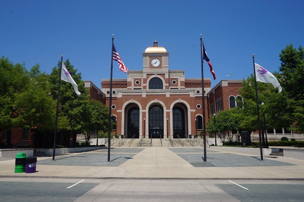 A large brick building in Lewisville, featuring a clock tower and three arched entrances, is surrounded by five flagpoles displaying flags. Green trees frame the scene under a clear blue sky. The foreground showcases a paved area leading to this impressive company headquarters.