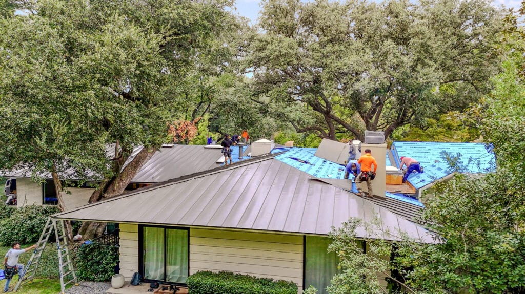 Roofers focus on crucial roofing repairs in a house surrounded by trees, installing blue underlayment and sleek metal roofing. Some use ladders, while others work on the roof in safety gear. The scene is bustling with construction materials all around.