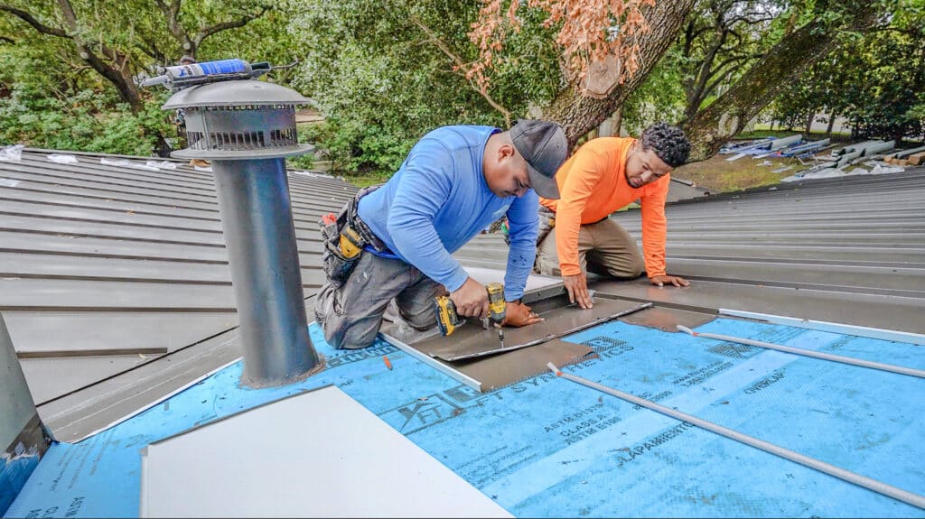 Two workers are skillfully installing a metal roof on a house, perfecting their craft. One expertly wields a drill while the other assists with positioning the panels for this meticulous metal roofing project, all amidst an array of trees and essential roofing materials.