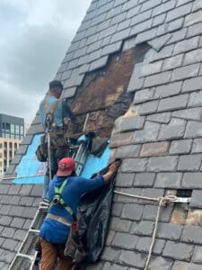 Two construction workers in safety gear are performing slate roofing repairs. One worker is on a ladder, while the other holds a piece of black tarp. The roof has a section removed, exposing wooden planks underneath. A building is visible in the background.