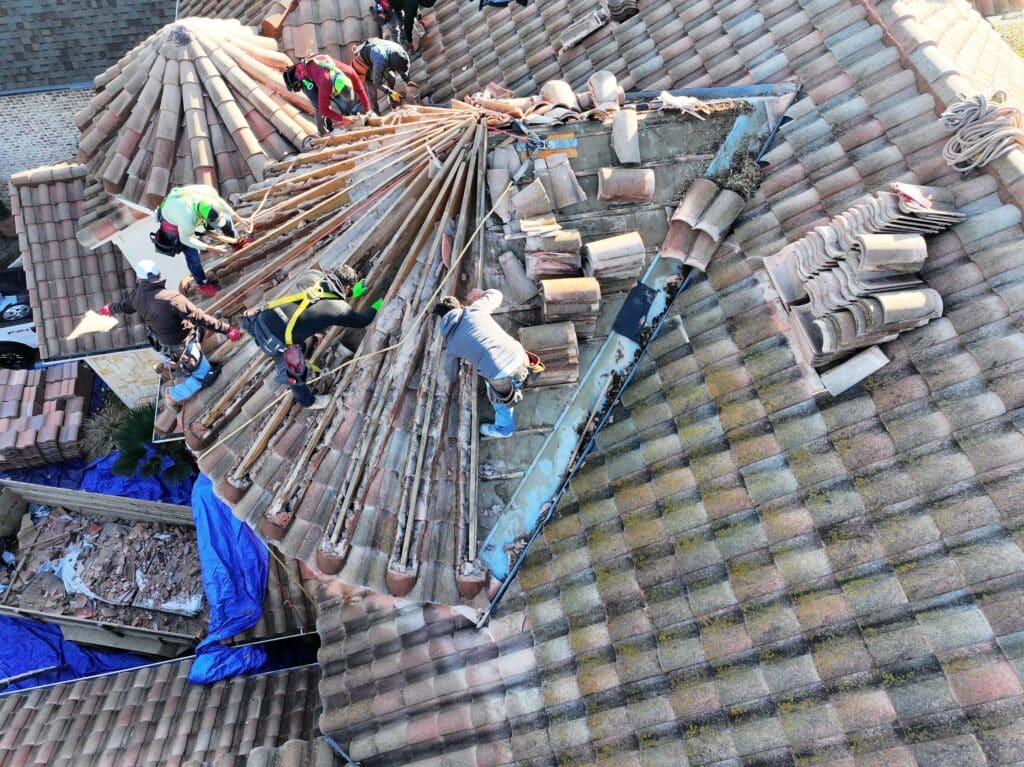 An aerial view captures construction workers engaged in roof repairs. Clad in safety harnesses and helmets, they maneuver among wooden beams and tiles. The rooftop showcases a blend of beige and brown tiles, with several neatly stacked nearby, ready for precise tile placement.