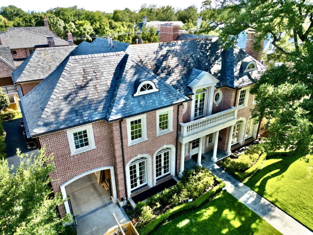 Aerial view of a large, elegant brick house with a dark slate roof crafted by a renowned roofing company in Dallas. It features multiple windows, a central columned entrance with an inviting balcony above, and meticulously manicured lawns, all nestled among trees and neighboring homes.