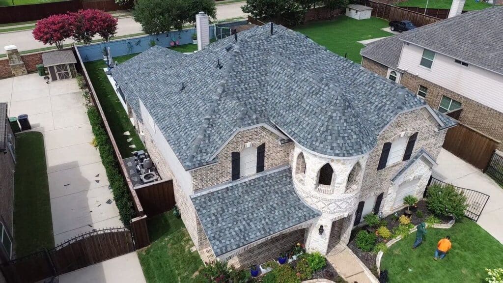 Aerial view of a two-story house with a gray shingled roof, brick exterior, arched entrance, and landscaped front yard in Dallas-Fort Worth. Two people are standing on the lawn, possibly discussing roof replacement with trusted experts.