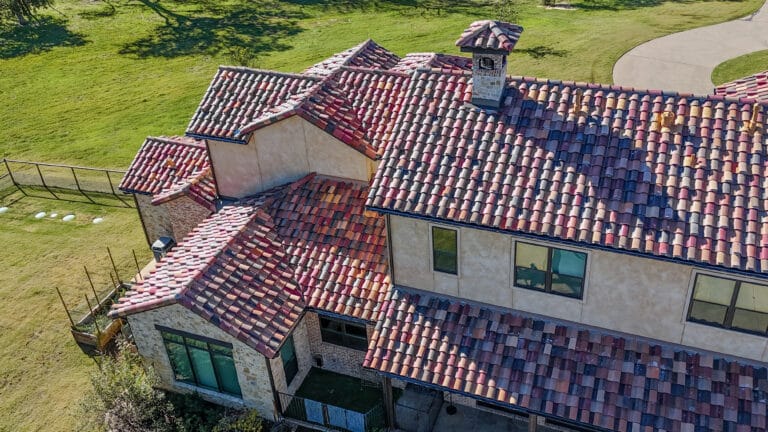 Aerial view of a house with a multi-level roof featuring red, pink, and purple ceramic tiles, beige stucco walls, and several large windows, surrounded by a grassy yard and a curved driveway.