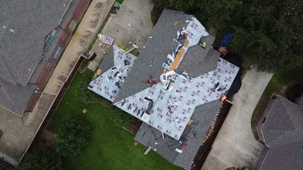 Aerial view of workers installing new roofing materials on a house. Sections of the roof are covered with protective sheeting, and tools and materials are scattered across the shingles. Nearby lawns and driveways are visible.