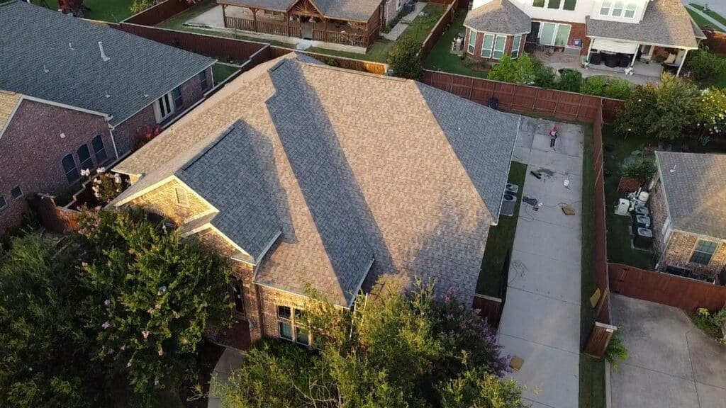 Aerial view of a suburban Dallas-Fort Worth house with a large driveway, fenced backyard, and trees. Trusted experts in roof replacement serve homes like these, with other houses and long shadows visible across the scene.