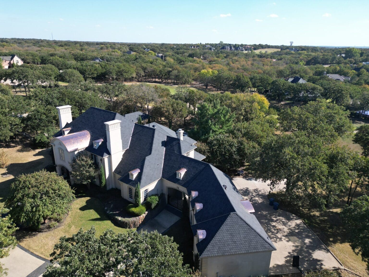 A large, light-colored house with a dark roof—recently updated by trusted experts—sits surrounded by trees and greenery in a spacious Dallas-Fort Worth neighborhood under a clear blue sky.