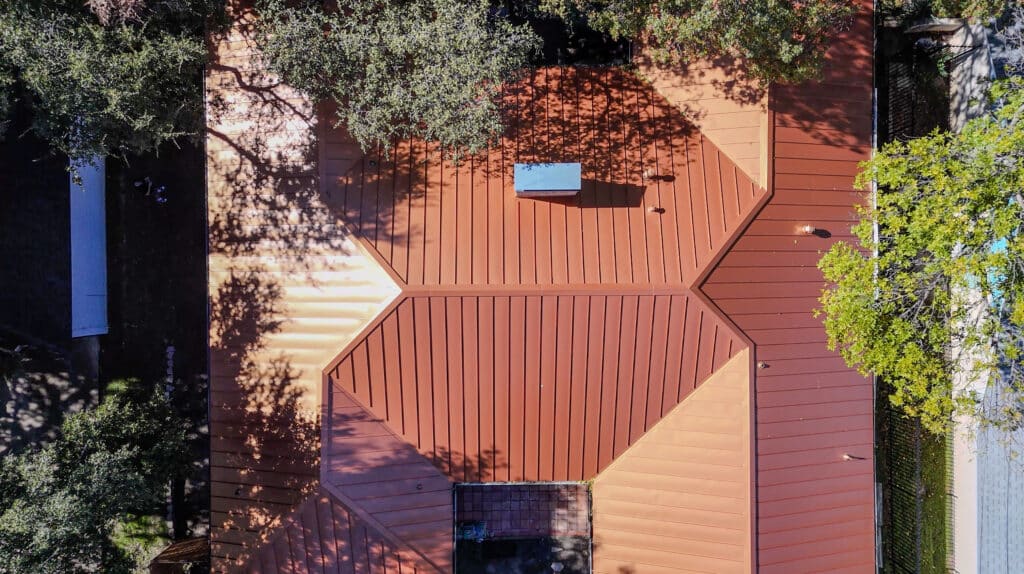 Aerial view of a red metal roofing with geometric sections, surrounded by trees casting shadows on the roof and ground. A small rectangular vent is visible on one section of the roof.