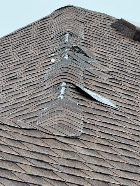 Damaged roof ridge with several shingles lifted and curled, exposing the underlayment—possible storm or wind damage. Trusted experts in DFW can assess and handle roof repairs, while the surrounding shingles appear intact.
