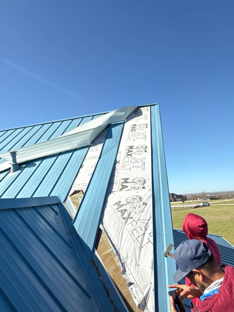 Two workers perform metal repairs and install insulation and siding on a slanted blue roof under a clear sky. The roof is partially covered with white synthetic underlayment labeled "MFT MAXFELT. - T Rock Roofing and Contracting Two workers perform metal repairs and install insulation and siding on a slanted blue roof under a clear sky. The roof is partially covered with white synthetic underlayment labeled "MFT MAXFELT.