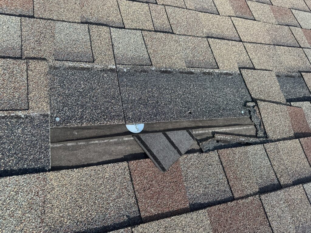 Close-up of a shingled roof in need of roof repairs, with missing and broken shingles exposing black underlayment and a metal nail—an issue trusted experts in DFW can address.