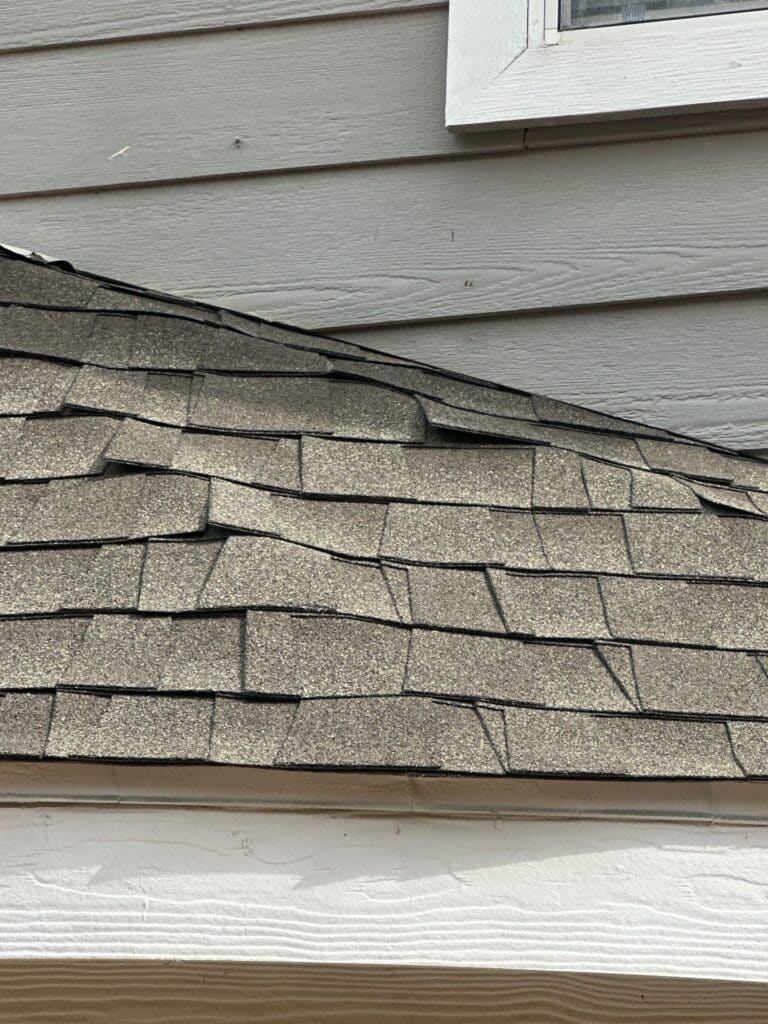 Close-up of a house roof in DFW shows gray asphalt shingles with some uneven edges; trusted experts recommend roof repairs. Gray horizontal siding and part of a window are visible above the roofline.