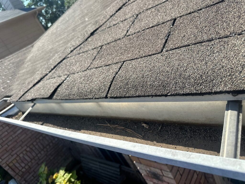 A close-up view of a house roof with asphalt shingles and an empty metal gutter, showing some dirt and debris inside the gutter—perfect for highlighting professional gutter services. Sunlight casts shadows on the roof and wall below. - T Rock Roofing and Contracting A close-up view of a house roof with asphalt shingles and an empty metal gutter, showing some dirt and debris inside the gutter—perfect for highlighting professional gutter services. Sunlight casts shadows on the roof and wall below.