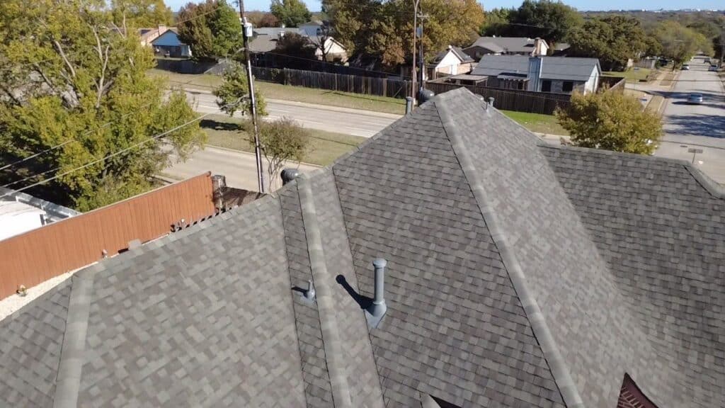 A view of a house’s gray shingle roof taken from above, showing roof vents and ridges. Located in a Dallas-Fort Worth suburb, the image highlights the work of Professional Roof Services with nearby trees, roads, and homes in the background.