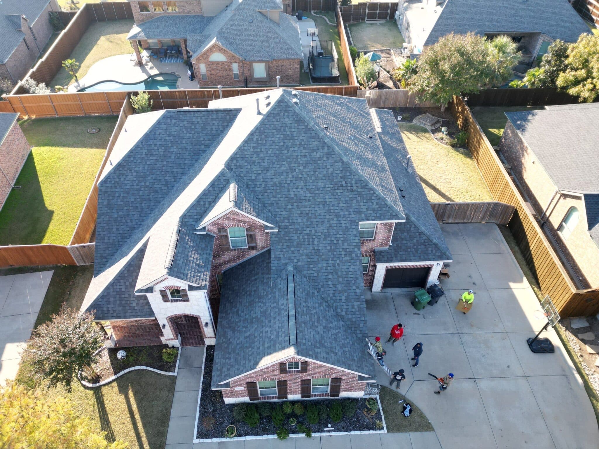 Aerial view of a two-story brick house with a gray roof, surrounded by a fenced backyard in Dallas-Fort Worth. Several people are gathered in the driveway, some standing and some sitting, near green bins—possibly discussing professional roofing services.
