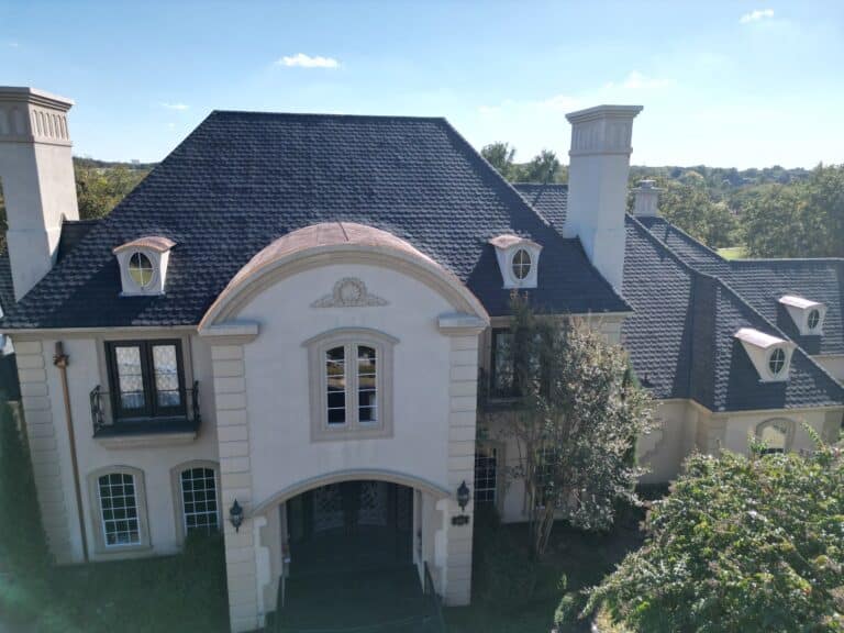 A large, elegant two-story house with a dark slate roof—recently updated by Professional Roof Replacement—features arched windows and decorative chimneys, surrounded by greenery and trees under a clear blue sky.