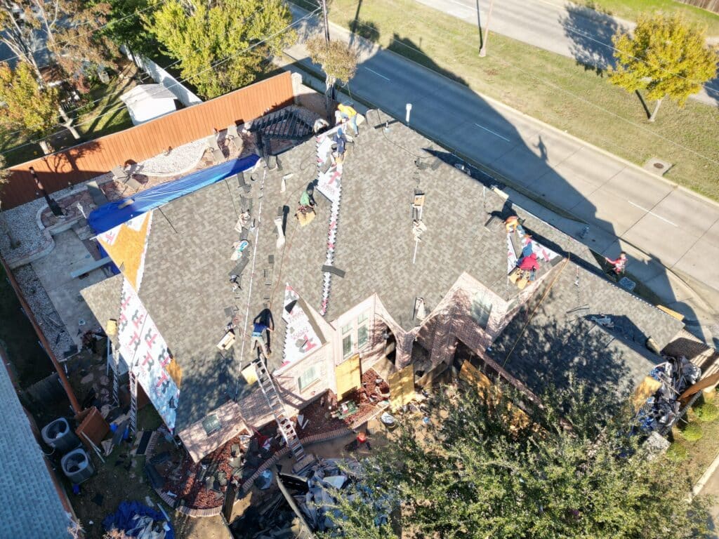 Aerial view of a house under construction or repair in Dallas-Fort Worth, with Professional Roof Services workers on the roof, building materials scattered around, and a blue tarp covering part of the structure. Trees and a street are visible nearby.