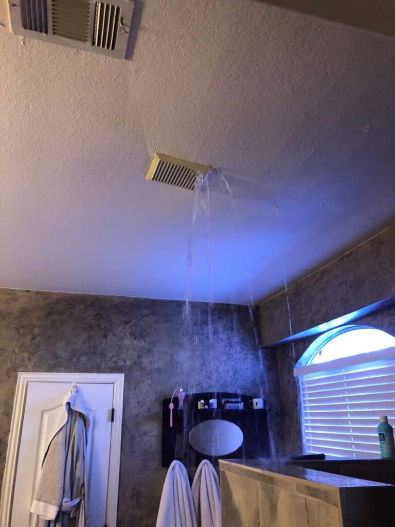 Water is pouring from a ceiling vent in a bathroom, likely due to frozen pipes or winter storm damage repair needs. Towels hang on hooks, and toiletries are visible on a shelf near a window with blinds.