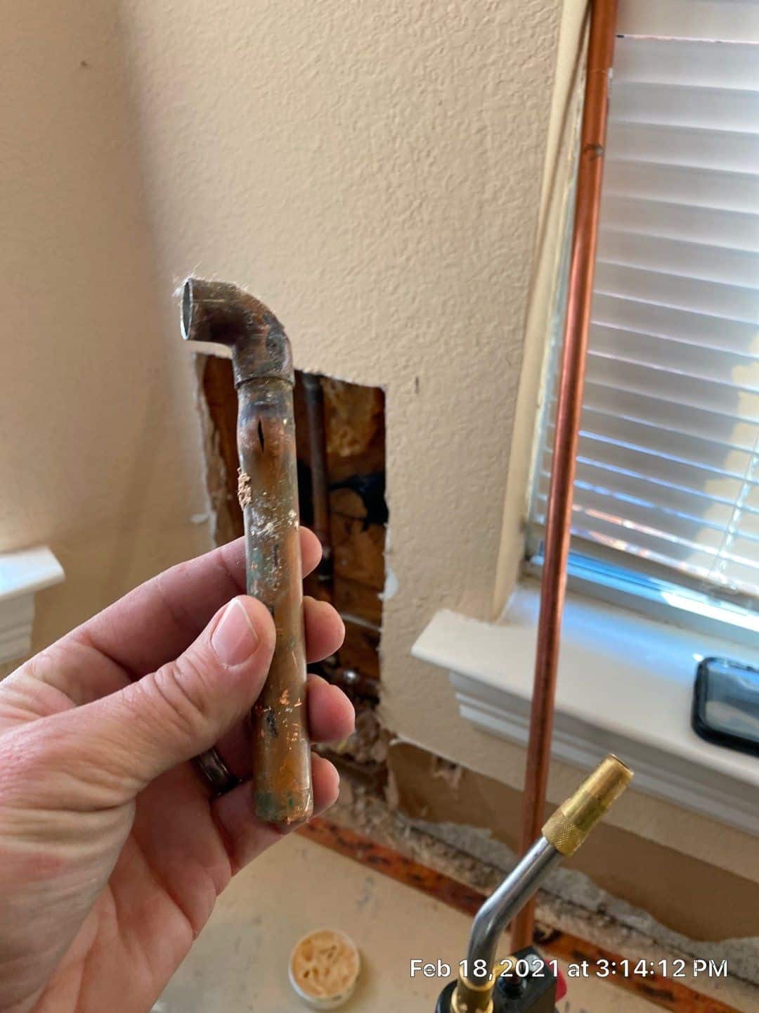 A hand holds a corroded, bent copper pipe in front of a wall with a rectangular hole exposing insulation—likely the result of frozen pipes. Tools and new copper piping are nearby, with a window and blinds on the right.