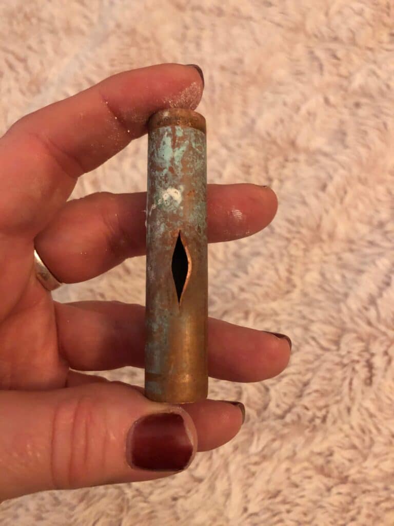 A hand holding a corroded copper pipe with a large vertical split down the side, likely from frozen pipes, set against a soft, beige textured background. The person's fingernails are painted dark red.