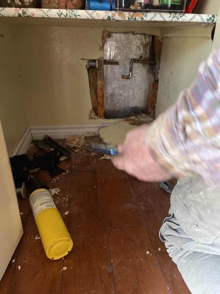A person kneels on a wooden floor, repairing a wall under a sink. The wall is open, exposing pipes—possibly dealing with frozen pipes—and tools including a yellow propane tank are scattered nearby. The person is holding a tool in their hand.