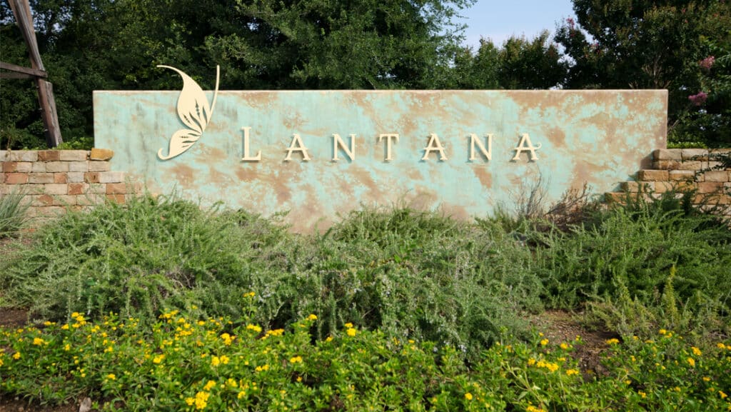 A large stone sign with the word "Lantana" and a butterfly graphic, surrounded by green shrubs and yellow flowers, with trees in the background.