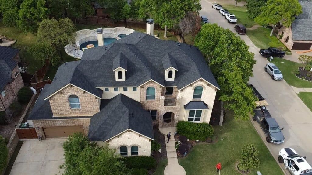 Aerial view of a large two-story house with a dark roof, light brick exterior, circular driveway, manicured lawn, and backyard pool. Several cars are parked along the street and driveway. Trees surround the property.