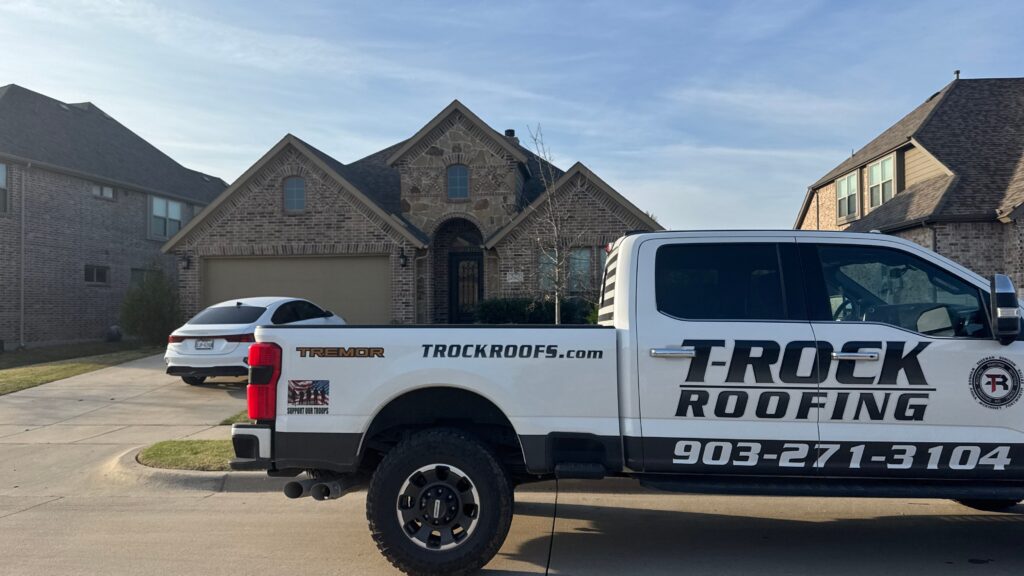 A white pickup truck with "Trock Roofing" branding is parked on a suburban street in front of a brick house with a driveway and another white car.