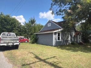 A small gray house with a brown roof sits beside a grassy yard. A white utility truck and a red car are parked nearby. Trees and blue sky with clouds are in the background.